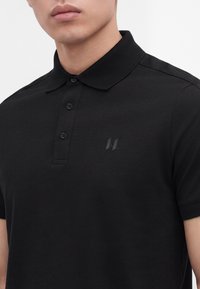 Homme portant un polo noir à manches courtes avec une patte de boutonnage à trois boutons et un petit logo embossé sur la poitrine.