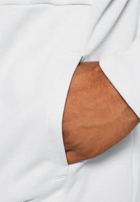 Main partiellement dans la poche latérale d'un vêtement blanc texturé, montrant les détails du tissu et la texture de la peau.