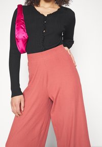Zwart geribbeld langemouwen shirt met een ruches halslijn, gecombineerd met roze wijde broek en een glanzende fuchsia handtas. Textuur en kleuren contrasteren.