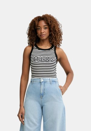 Donna con capelli ricci che indossa un top senza maniche a strisce bianche e nere e jeans larghi azzurri a vita alta, in piedi con una mano in tasca.