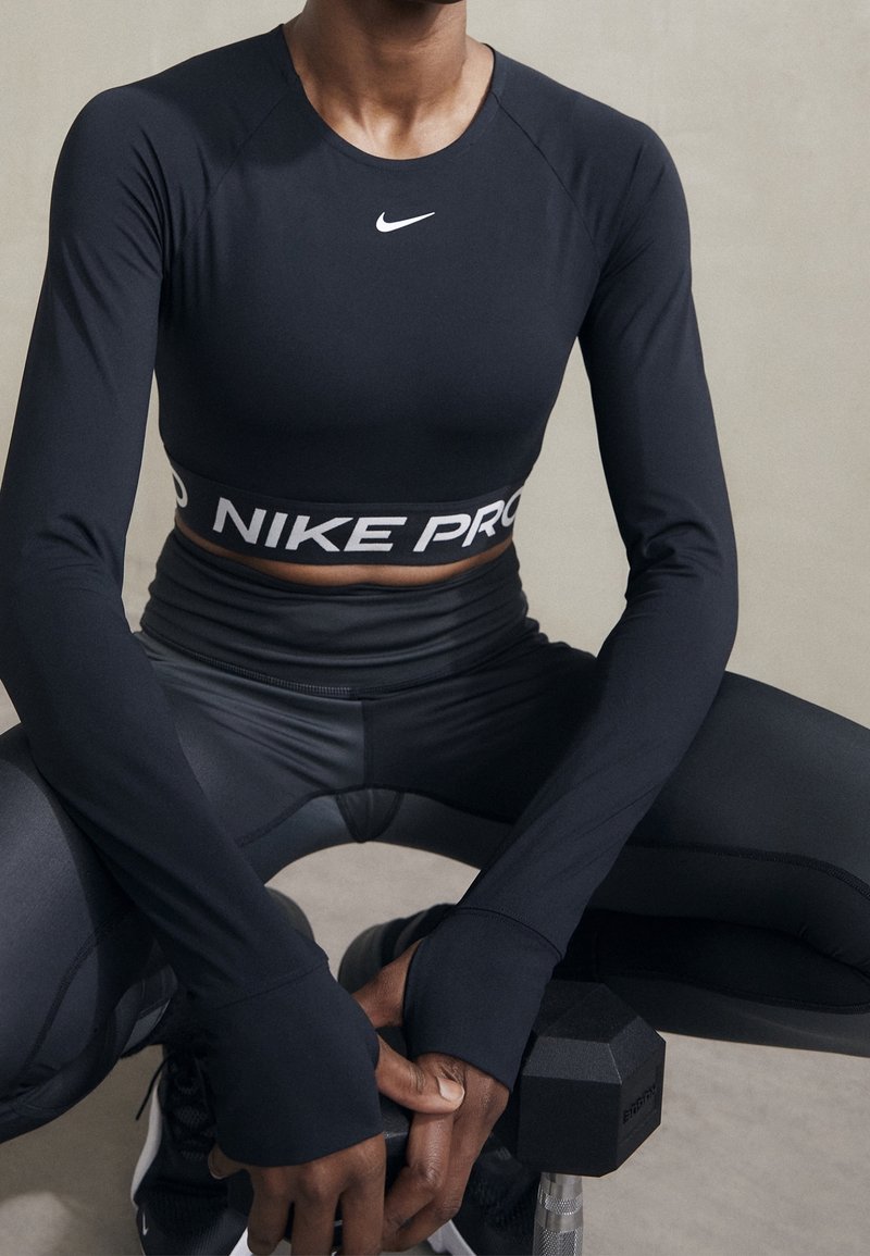 Henkilö mustissa Nike Pro -urheiluvaatteissa kyykkää, kädet ristissä mustan käsipainon ympärillä yksinkertaisella taustalla.