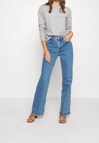 Felpa grigia con la scritta "Calvin Klein", abbinata a jeans a vita alta, larghi di colore azzurro chiaro, e sandali a scivolo marroni su uno sfondo neutro.
