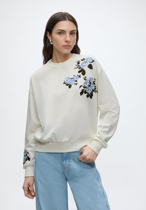 Vrouw met donker haar die een witte trui draagt met blauwe bloemenborduursel op de schouder en mouw, gecombineerd met lichtblauwe spijkerbroek.