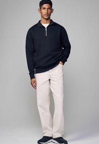 Zwarte zip-up sweatshirt met lange mouwen, gecombineerd met lichtbeige broeken en zwarte sneakers. De sweatshirt heeft een staande kraag.
