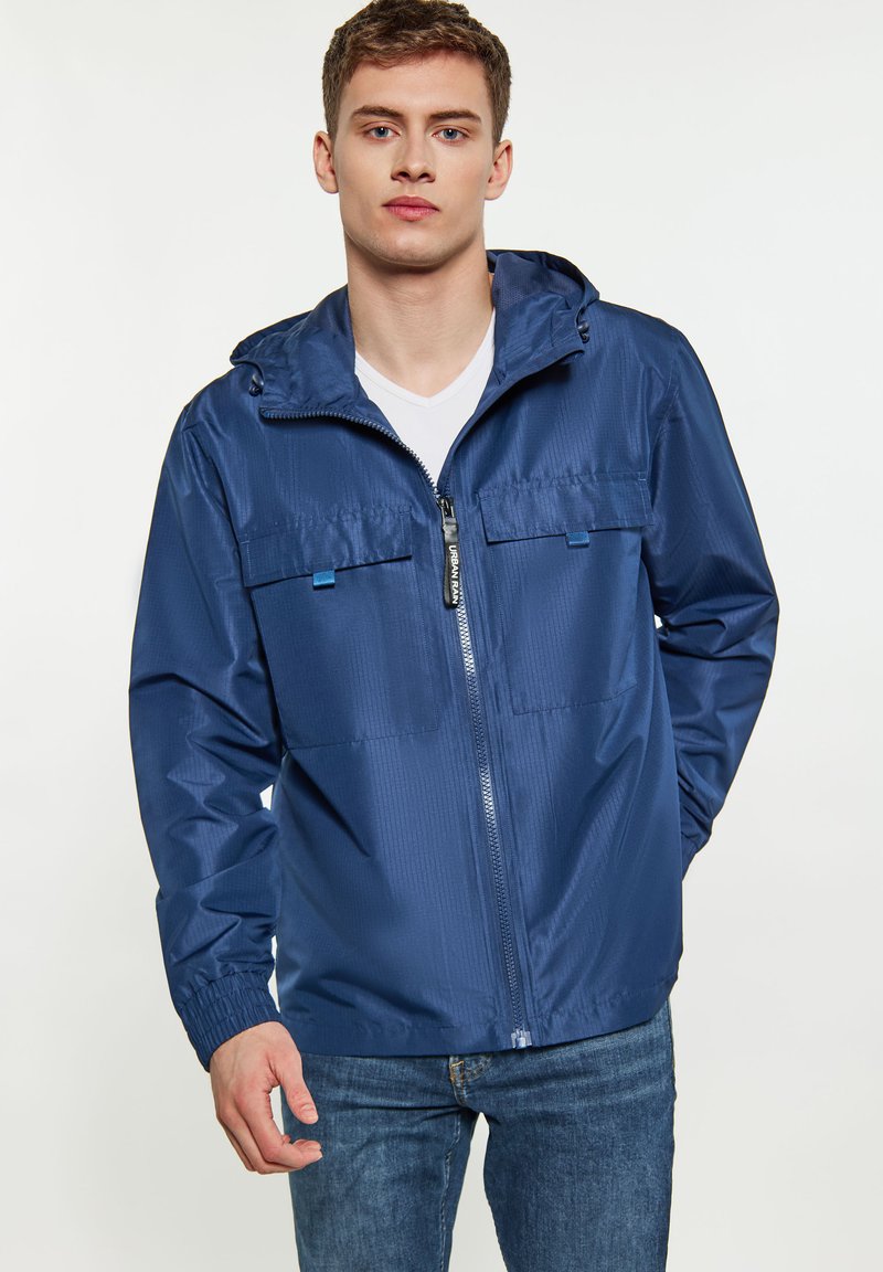 Tamsiai mėlynas vėjas neperpučiamas švarkas, pagamintas iš lengvos, tekstūruotos medžiagos. Yra priekinis užtrauktukas, dvi krūtinės kišenės ir gobtuvas.