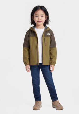 Małe dziecko stojące w oliwkowo-brązowej kurtce z kapturem, białej koszulce, niebieskich dżinsach i beżowych trampkach na jednolitym tle.