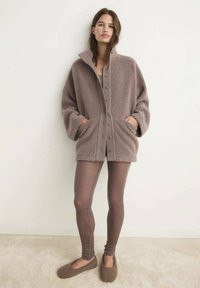 Veste en polaire marron oversize avec des boutons et des poches, portée sur des leggings marron assortis et des chaussures à enfiler confortables. Texture douce.