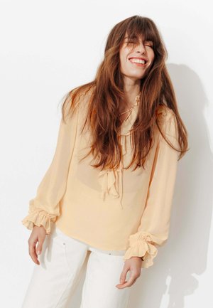 Jeune femme aux longs cheveux bruns portant un chemisier pêche à volants et un pantalon blanc, souriant les yeux fermés sur un fond blanc.