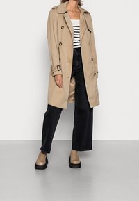 Beżowy trencz z dwurzędowym zapięciem, dużymi guzikami i paskiem. Noszony na paskowanym topie, zestawiony z ciemnymi szerokimi spodniami jeansowymi i beżowymi butami.
