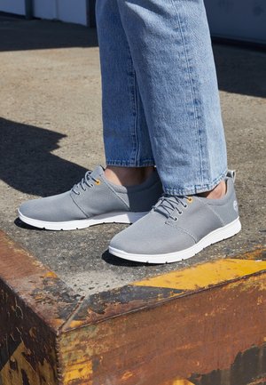 Osoba nosząca jasnoniebieskie dżinsy i szare siateczkowe trampki stojąca na zardzewiałej metalowej krawędzi na betonowej powierzchni.