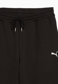 Juodos kelnės, pagamintos iš švelnios medžiagos, su elastine juosta, reguliuojama raišteliu ir baltu Puma logotipu apatinėje dešinėje pusėje.