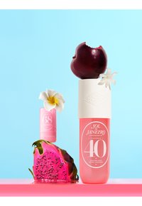 Dwie różowe buteleczki perfum Sol de Janeiro z ugryzioną śliwką, owocem smoka i białymi kwiatami na niebieskim tle i różowej powierzchni.