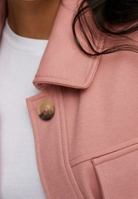 Różowa, fakturowana kurtka z dużymi klapami. Zdobiona okrągłym, błyszczącym guzikiem. W tle widoczna biała koszula.