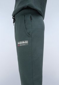 Dunkelgrüne Sweatpants aus weichem Material mit Seitentaschen, einem elastischen Bund und einem großen Logopatch am Oberschenkel.