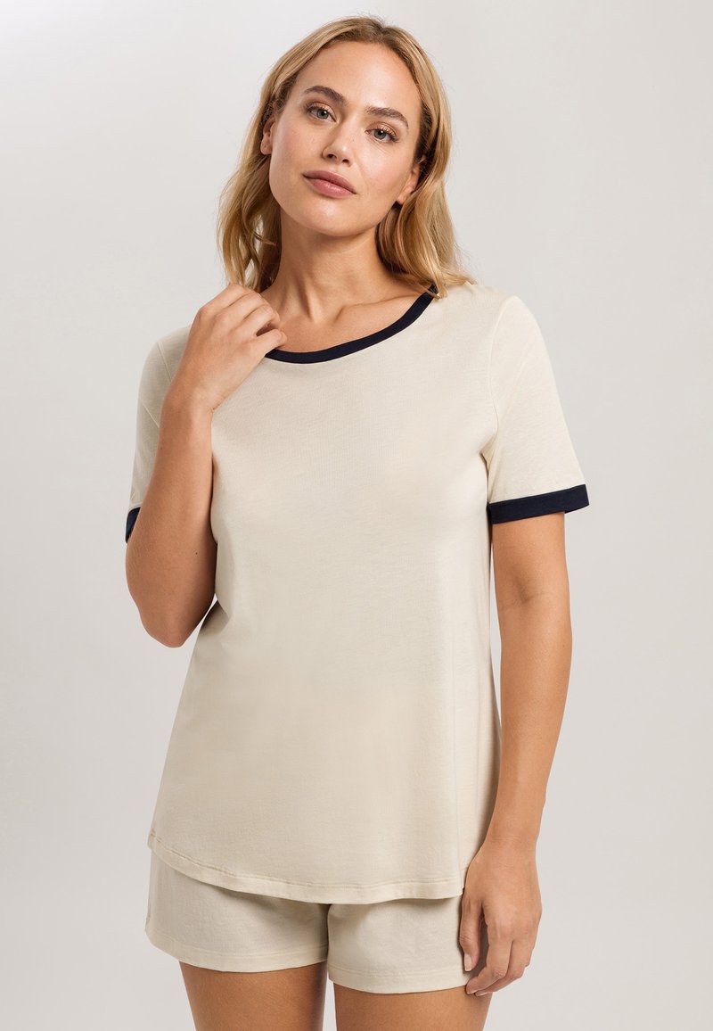 Beige top s krátkým rukávem s námořnickým lemováním kolem výstřihu a rukávů, v kombinaci s odpovídajícími béžovými šortkami vyrobenými z měkké tkaniny s hladkou strukturou.