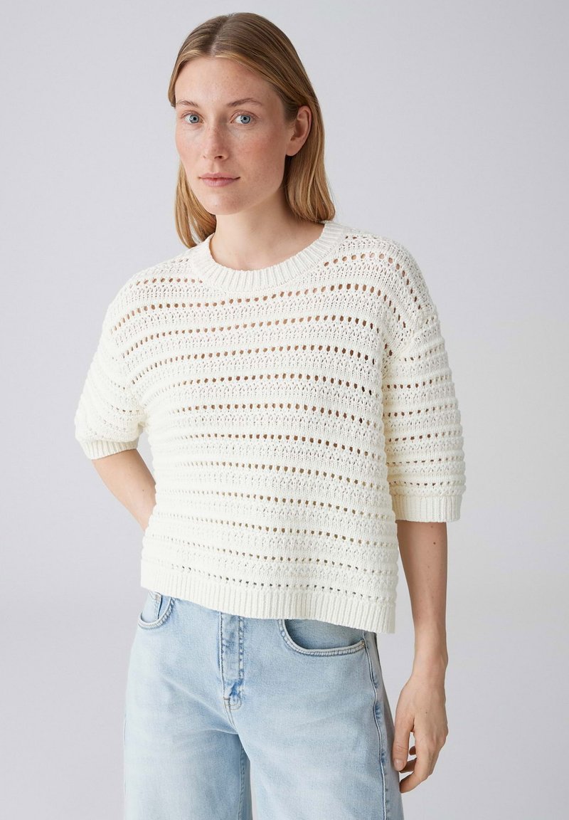 Frau trägt weißen Kurzarm-Pullover aus Häkelstrick und hellblaue Jeans, steht mit der Hand in der Hüfte vor einem einfarbigen Hintergrund.