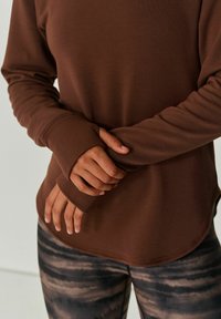 Bruine langemouwen top met een ronde zoom, voorzien van duimgaten in de manchetten, gecombineerd met patroonleggings in grijs- en bruintinten.