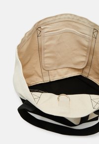 Sac en toile avec un intérieur non doublé, comportant une poche, un fond noir et un anneau en métal pour fixation. Schéma de couleurs noir et crème.