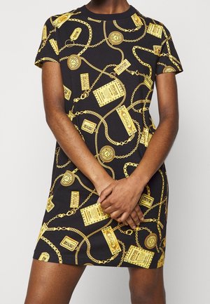 Person trägt ein schwarzes kurzärmeliges Kleid mit Muster aus goldenen Ketten und rechteckigen goldenen Anhängerdesigns, steht mit verschränkten Händen vorne.