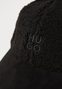 Črna kapa z vložkom iz materiala podobnega flisu na vrhu, ki ima embosirano logo z napisom "HUGO." Vizir je gladek in rahlo ukrivljen.