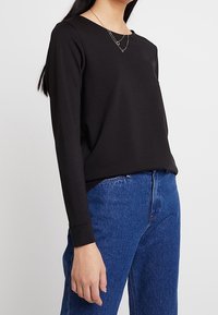 Schwarzes Langarmtop aus glattem Stoff mit rundem Halsausschnitt. Kombiniert mit hochgeschnittenen blauen Jeans. Einfaches Design.