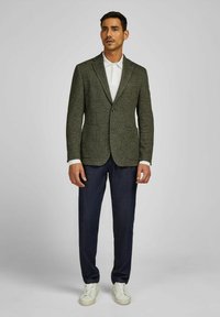 Man met een donkergroene gestructureerde blazer, een wit polo-shirt, donkerblauwe broeken en witte sneakers, staande tegen een effen lichte achtergrond.