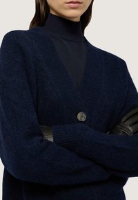 Námořnický pletený cardigan s výstřihem do V, který má jediné zapínání na knoflík a texturovanou tkaninu. Pod ním je vidět přiléhavý černý rolák.