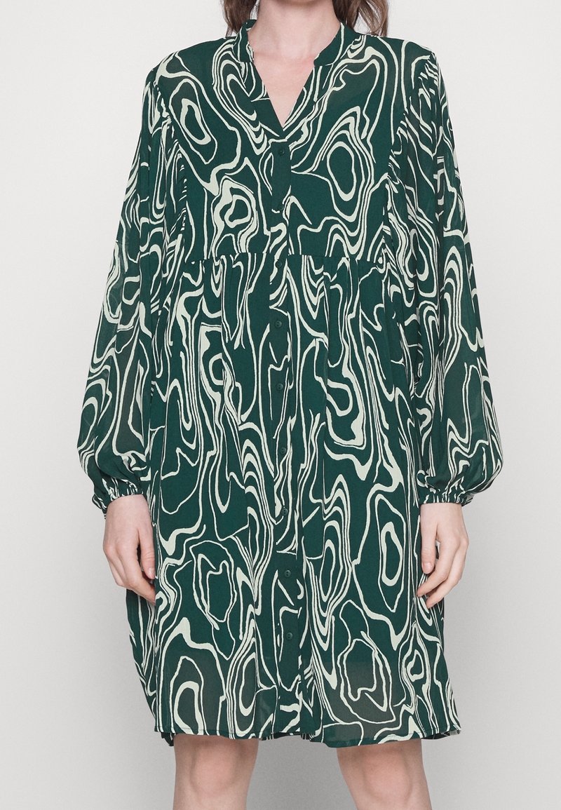 Robe verte à manches longues avec motif tourbillonnant blanc abstrait, boutonnée sur le devant et taille froncée, portée par une personne debout.
