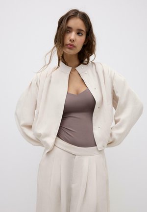 Kvinde med langt hår iført en løs, off-white jakke, taupefarvet tætsiddende top og matchende bredbenede plisserede bukser, stående med hænderne på hofterne.