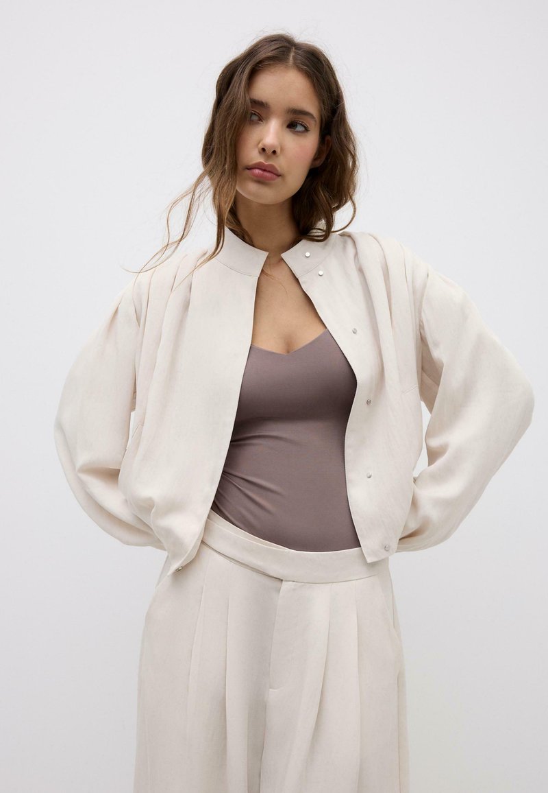 Femme aux cheveux longs portant une veste ample écrue, un haut taupe ajusté et un pantalon large plissé assorti, debout avec les mains sur les hanches.