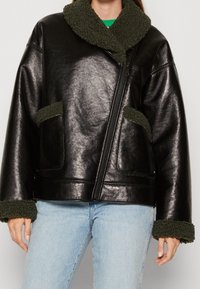 Veste en cuir noir avec un col et des poignets en peluche verte, dotée de deux poches avant et d'une fermeture éclair. Texture lisse avec une finition brillante.