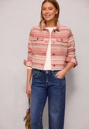 Uśmiechnięta kobieta, w czerwono-białej wzorzystej krótkiej kurtce na białej koszuli, niebieskich dżinsach, trzymająca brązową torebkę z frędzlami.