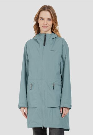 Woman wearing a light blue hooded raincoat with front zipper and zippered pockets, standing against a plain gray background.