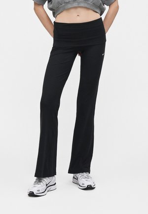 Persoon draagt zwarte uitlopende legging en witte Nike sneakers, met handen achter de rug en grijze cropped top die de buik laat zien.