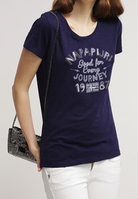 Camiseta azul marino con cuello redondo, que presenta el texto gráfico plateado "NAPAPIJRI Good for Every Journey 1987". Combinada con un bolso negro con lentejuelas.