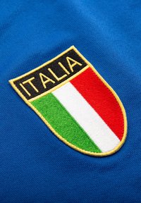 Schildförmiger Aufnäher mit den Farben der italienischen Flagge Grün, Weiß und Rot und dem Wort "ITALIA" in Gelb auf Schwarz, auf blauem Stoff aufgenäht.