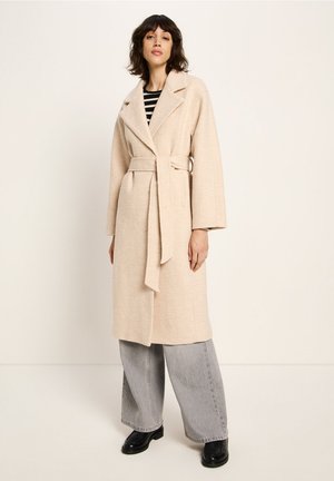 Beige Wickelmantel mit einem Gürtel und weiten Ärmeln, getragen über einem schwarz-weiß gestreiften Oberteil. Das Modell kombiniert ihn mit grauen, weiten Hosen und schwarzen Schuhen.
