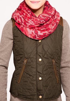 Femme portant une écharpe infinie rouge à motifs, un gilet matelassé vert olive avec des boutons dorés, et un haut beige à manches longues.