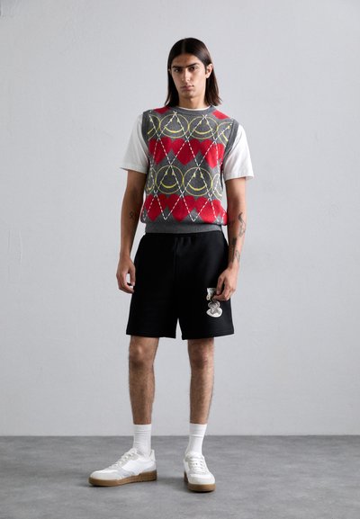 Grå genser vest med røde og gule geometriske mønstre over en hvit t-skjorte, kombinert med svarte shorts og hvite joggesko med brune såler.