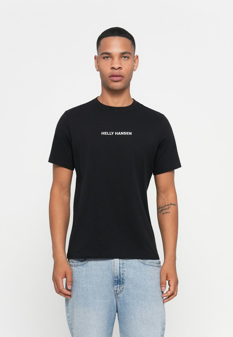 Man with short hair wearing black Helly Hansen t-shirt and light blue jeans, standing against a plain white background.