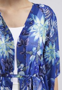 Žena oblečená v modrém květinovém průhledném kimono-stylovém oděvu svázaném v pase přes bílý top.