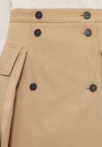 Jupe beige taille haute avec des boutons noirs disposés en deux rangées verticales et une poche latérale avec rabat.
