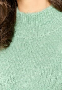 Svetlozelena pletena pulover z rebrastim visokim ovratnikom. Mehka tekstura z vodoravnimi in navpičnimi pleteninskimi vzorci. Brez vidnih kovinskih delov ali okraskov.