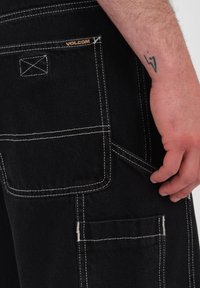 Czarne dżinsowe spodnie z białymi szwami, wieloma kieszeniami i logo Volcom. Ręka znajduje się przy kieszeni do regulacji po bokach.