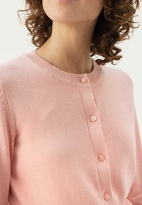 Light pink, knitted cardigan with a round neck, featuring four buttons down the front and ribbed cuffs, made from soft textile material.