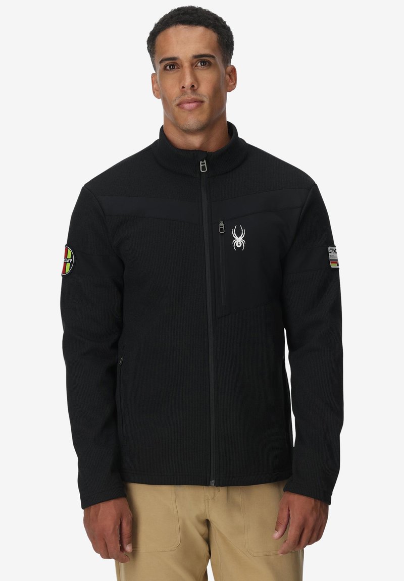 Giacca in pile nera con colletto alto, dotata di zip frontale, dettagli con logo sul petto e sulla manica, e un design in tessuto testurizzato.