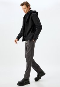 Chaqueta softshell negra con capucha, cierre frontal y bolsillos en el pecho; combinada con pantalones cargo grises y botas negras con suela gruesa.