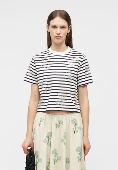 Γυναίκα που φοράει άσπρο και ναυτικό μπλε ριγέ κοντομάνικο τοπ με floral σχέδια και κρεμ φούστα με πράσινη floral κεντήματα, κρατώντας μια μαύρη τσάντα.