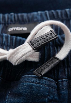 Pantaloni in denim di un blu scuro, con coulisse bianca e etichette nere con marchio, che mettono in risalto i dettagli della texture e delle cuciture.