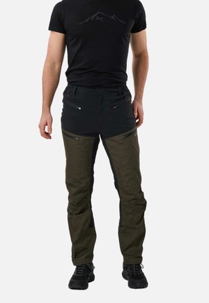 Mężczyzna ubrany w czarne i oliwkowe outdoorowe spodnie na zamek, czarne buty trekkingowe i czarną koszulkę z logo góry.